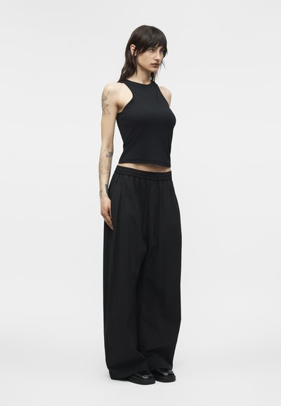 Γυναίκα με σκουρόχρωμα μαλλιά φοράει μαύρο αμάνικο crop top, μαύρο παντελόνι με φαρδιά πόδια, μαύρα παπούτσια και έχει τατουάζ στα χέρια και τον ώμο.