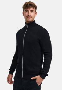 Musta vetoketjullinen paita, jossa korkeakaulus, teksturoitu neulekuvio ja kontrastivärinen vetoketju. Resorit hihansuissa ja helmassa.