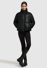 Kvinde iført en sort puffjakke, sorte leggings og sorte knæhøje støvler, stående med hænderne i lommen mod en ensfarvet baggrund.