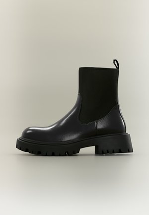 Bottine en cuir noir avec semelle épaisse en caoutchouc, panneau latéral élastique et languette à l’arrière pour un enfilage facile.