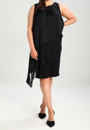 Frau trägt ein ärmelloses schwarzes asymmetrisches Midikleid mit einer mehrlagigen, strukturierten Überlagerung und schwarzen spitzen Pumps, steht mit einer Hand hinter dem Rücken.