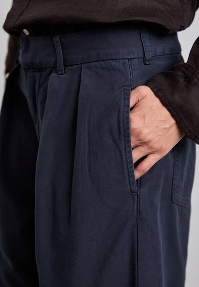 Mörkblå byxor med en slät textur, som har frontveck och sidofickor. Midjan har bälteshällor och en skarp sömdetalj.