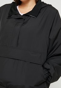 Juodas, lengvas striukė su užtrauktuku ant apykaklės, elastingomis rankovėmis ir laisvu kirpimu, turinti priekinę kišenę. Lygi audinio tekstūra.