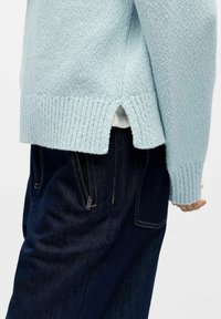 Lyseblå strikket genser med ribbede mansjetter og kant, med en sidesplitt. Kombinert med mørk denimjeans med glidelåslommer.