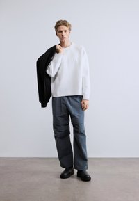 Λευκό φούτερ με υφή στην μπροστινή πλευρά, γκρι παντελόνι cargo με πολλαπλές τσέπες και μαύρες αστραγάλες μποτάκια, στέκεται μπροστά σε απλό φόντο.