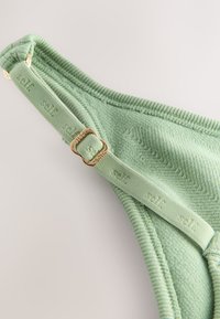 Rohelise tekstuuriga puuvillane rinnahoidja rihm, millel on reguleeritav kuldse tooniga metallosa ja reljeefne bränding; riidekattel on kalasaba muster.