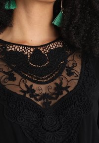 Top negro con intrincados bordados de encaje negro en el escote, patrones florales y un forro de malla transparente. Pendientes de borlas verdes visibles.