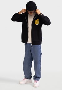 Čierna mikina na zips so žltou grafikou na hrudi, béžové tričko pod ňou, voľné modré džínsy a biele tenisky s ružovými šnúrkami.