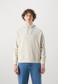 Beige sweatshirt i mjukt tyg, med en framficka, dragsko i huvan och ribbade ärmslut. Matchas med blå denimjeans.