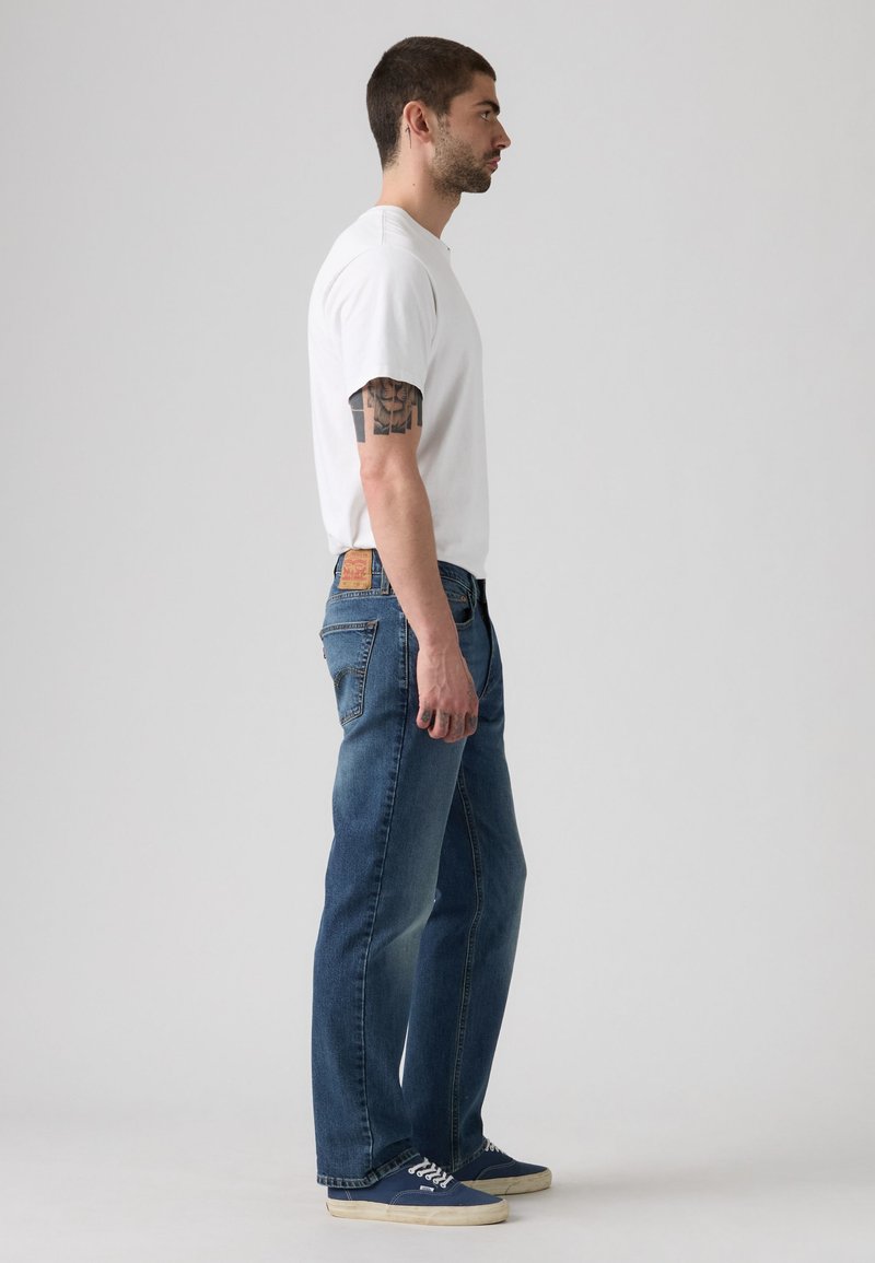 Man with tattoo wearing white t-shirt, blue jeans, and navy sneakers standing in profile against plain background.