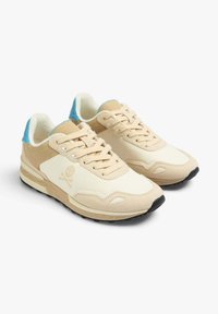 Beige sneakers with a beige leather and suede upper, light blue accents on the heel, textured rubber sole, and a skull logo on the side.