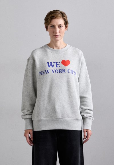 Siva pamučna majica s rebrastim manšetama i rubom. Na sebi ima crveno srce i plavi tekst s natpisom "VOLIMO NEW YORK CITY." Dizajn opuštenog kroja.