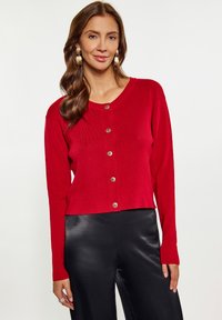Rode button-up cardigan met een ronde hals en lange mouwen. Voorzien van metalen knopen en een cropped zoom, gecombineerd met zwarte broeken.