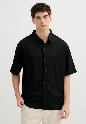 Joven con cabello rizado oscuro que lleva una camisa negra de manga corta abotonada y pantalones beige claros, de pie frente a un fondo blanco liso.