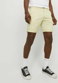 Shorts jaunes clairs avec revers, en coton, dotés d'une fermeture à bouton sur le devant et de poches latérales, associés à des baskets noires et des chaussettes blanches.