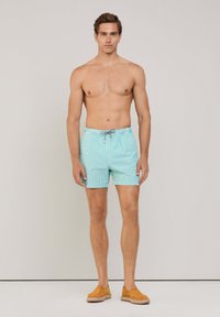 Costumi da bagno a righe blu chiaro con vita elastica e cordoncino, realizzati in tessuto che asciuga rapidamente. Il modello indossa delle scarpe slip-on color sabbia.