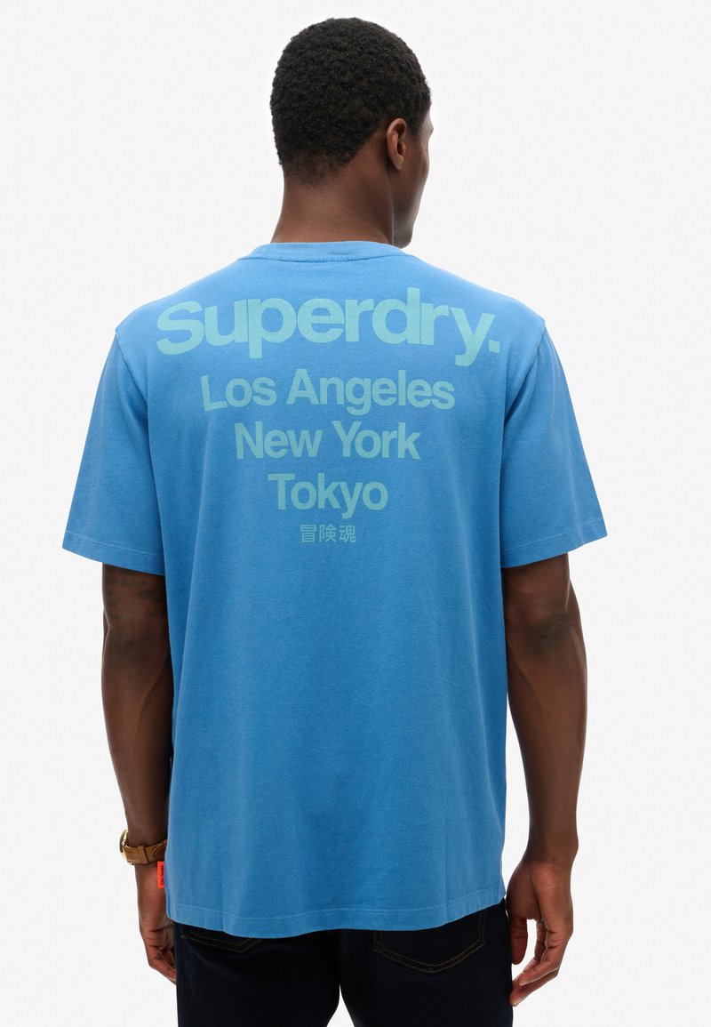 Blå bomulds t-shirt med stor hvid tekst på ryggen: "Superdry. Los Angeles New York Tokyo," med en afslappet pasform og korte ærmer.