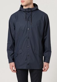 Una persona indossa una giacca da pioggia blu navy con cappuccio, caratterizzata da bottoni e tasche, su uno sfondo neutro chiaro.