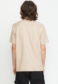 Beige kortærmet skjorte med lige snit i kanten, subtil tekstur og detalje med rygsøm. Har en enkelt løkke ved halsen.