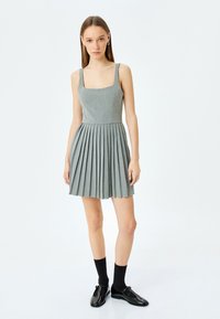 Robe grise plissée avec un décolleté carré et des bretelles larges. Portée avec des chaussettes noires et des chaussures. Tissu lisse avec une texture subtile.