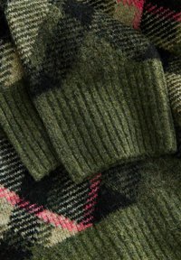 Close-up of a green wool sweater sleeve with ribbed cuff over a plaid wool fabric in green, beige, black, and red tones.