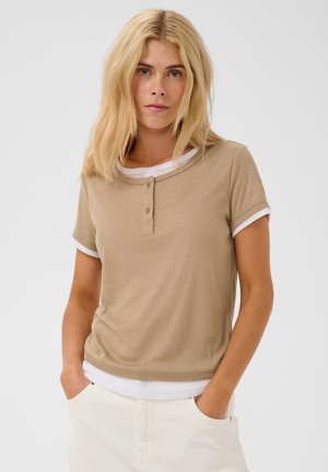 Femme aux longs cheveux blonds portant un t-shirt henley beige à manches courtes sur un haut blanc et un pantalon clair, debout les mains dans les poches.