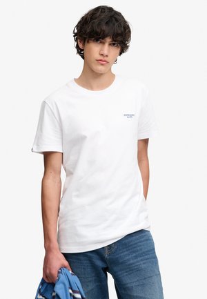 Młody mężczyzna z ciemnymi kręconymi włosami, ubrany w białą koszulkę Superdry i niebieskie dżinsy, trzymający w lewej ręce niebieską koszulę w paski.