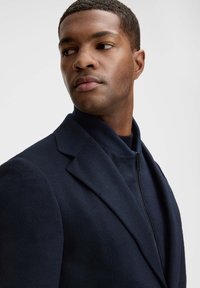 Granatowa marynarka z mieszanki wełny, z wysokim kołnierzem, zamkiem błyskawicznym z przodu i usztywnianymi ramionami. Gładka faktura, dopasowany krój, minimalistyczny design.