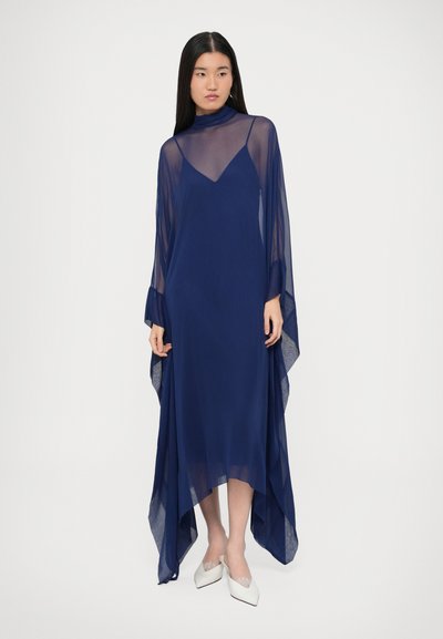 Rochie bleumarin din material transparent, cu guleraș înalt, mâneci lungi și tiv asimetric, fluid. Completată de pantofi albi cu vârf ascuțit.