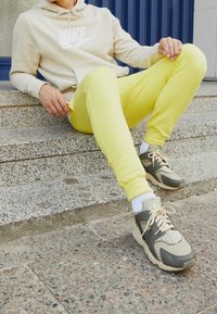 Osoba v béžové mikině Nike a jasně žlutých teplácích sedí na betonových schodech, má na sobě šedé a béžové tenisky s bílými ponožkami.