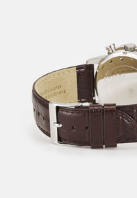 Bracelet de montre en cuir marron avec des détails texturés, fermoir en métal argenté et doublure intérieure lisse. Boîtier de montre visible en arrière-plan.