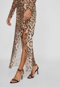 Kvinna i en leopardmönstrad lång klänning med en slits framtill, matchad med mörkbruna högklackade sandaler, ben och händer synliga.