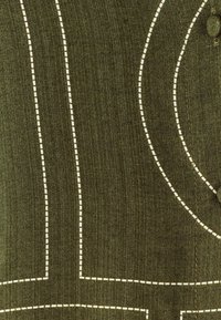 Olivgrüner Stoff mit kontrastierenden weißen Nähten. Enthält gewellte und gerade Linien, die ein strukturiertes Muster erzeugen. Beinhaltet ein Knopfdetail.