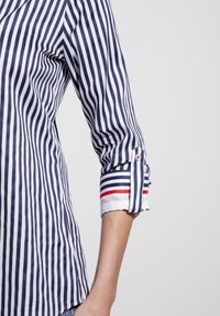 Navy and white striped shirt with rolled sleeves, featuring a button closure. Cuffs have red and blue accent stripes. Lightweight fabric.