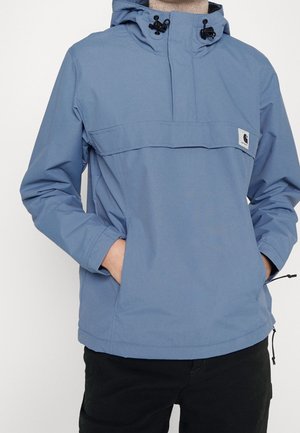 Veste à capuche bleu clair avec poches avant et cordons noirs sur la capuche, portée avec un pantalon noir.