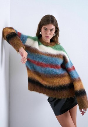 Ung kvinde læner sig op ad en hvid væg iført en blød, flerfarvet stribet sweater og en kort sort nederdel.