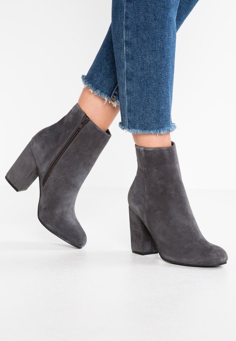 Jambes féminines portant des bottines en daim gris à talons blocs et zips latéraux, associées à un jean bleu à ourlet effiloché.