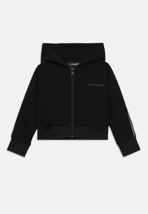 Black cropped hoodie made of soft fabric. Features a front zipper, hood, and ribbed hem. Left sleeve has decorative accent details.
