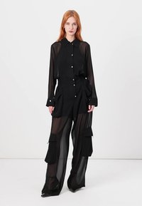 Crna prozirna košulja na dugmad u kombinaciji s hlačama širokih nogavica koje imaju cargo džepove, izrađene od lagane tkanine s glatkom teksturom.