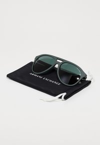 Ochelari de soare verzi din plastic cu lentile întunecate, având un design unghiular. Însoțiți de un sac negru cu șnur pe care este imprimat "ARMANI EXCHANGE".