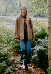 Tan fleecejakke med store lommer, parret med en sort leopardmønstret top og blå brede jeans. Sorte ankelstøvler fuldender outfittet.