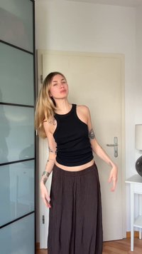 Νεαρή γυναίκα με μακριά ξανθά μαλλιά, φοράει μαύρο αμάνικο τοπ και φαρδιά καφέ παντελόνια, στέκεται σε ένα δωμάτιο με κλειστή πόρτα και έπιπλα.