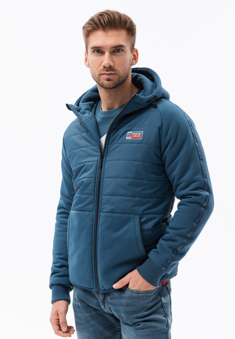 Giacca imbottita blu con cappuccio, dotata di chiusura con zip e tasche anteriori. Tessuto strutturato con una finitura liscia e dettagli del logo sulla manica.