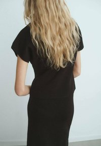 Vestido negro con mangas cortas, silueta ajustada y textura lisa. El cabello es largo y ondulado, peinado sobre el hombro. Fondo claro.