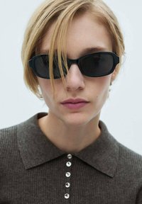 Sorte, rektangulære solbriller med mørke linser og tykke stel. Båret af en person med kort hår, sammen med en kulfarvet skjorte med krave.