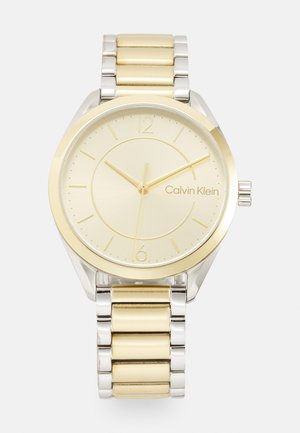 Reloj en tonos dorado y plateado con una esfera redonda, marcadores de horas minimalistas y una esfera cepillada, que cuenta con una pulsera de eslabones metálicos.