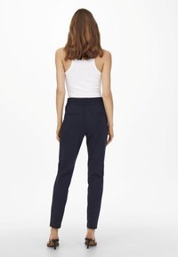 Marineblaue enganliegende Hose mit elastischem Bund, seitlichen Taschen und schmalem Schnitt, kombiniert mit einem weißen Tanktop und schwarzen High Heels.