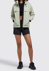 Jasnozielona kurtka wiatrówka z dwoma kieszeniami na piersi, regulowanym kapturem i przednim zamkiem błyskawicznym, zestawiona z czarnymi szortami i trampkami. Gładka faktura.