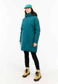 Chaqueta aislante de color verde azulado, diseño largo y recto con un cuello alto. Usada sobre pantalones negros y botas marrones con cordones negros y forro de piel.