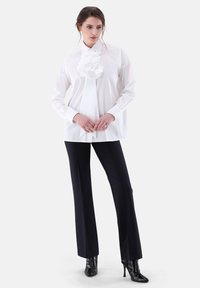 Weißes, übergroßes Hemd mit einer großen Stoffblüte am Kragen, kombiniert mit schwarzen, taillierten Hosen und hochhackigen Stiefeletten.